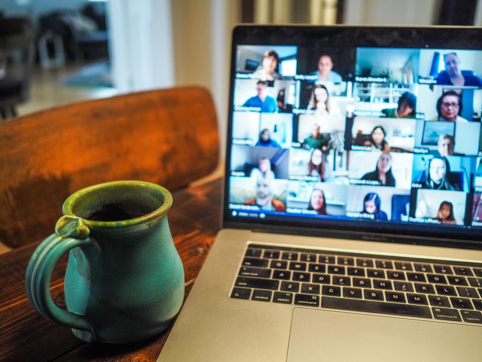 Schermo aperto di un pc con una videoconferenza, molte immagini di persone collegate. Sul tavolo una tazza.