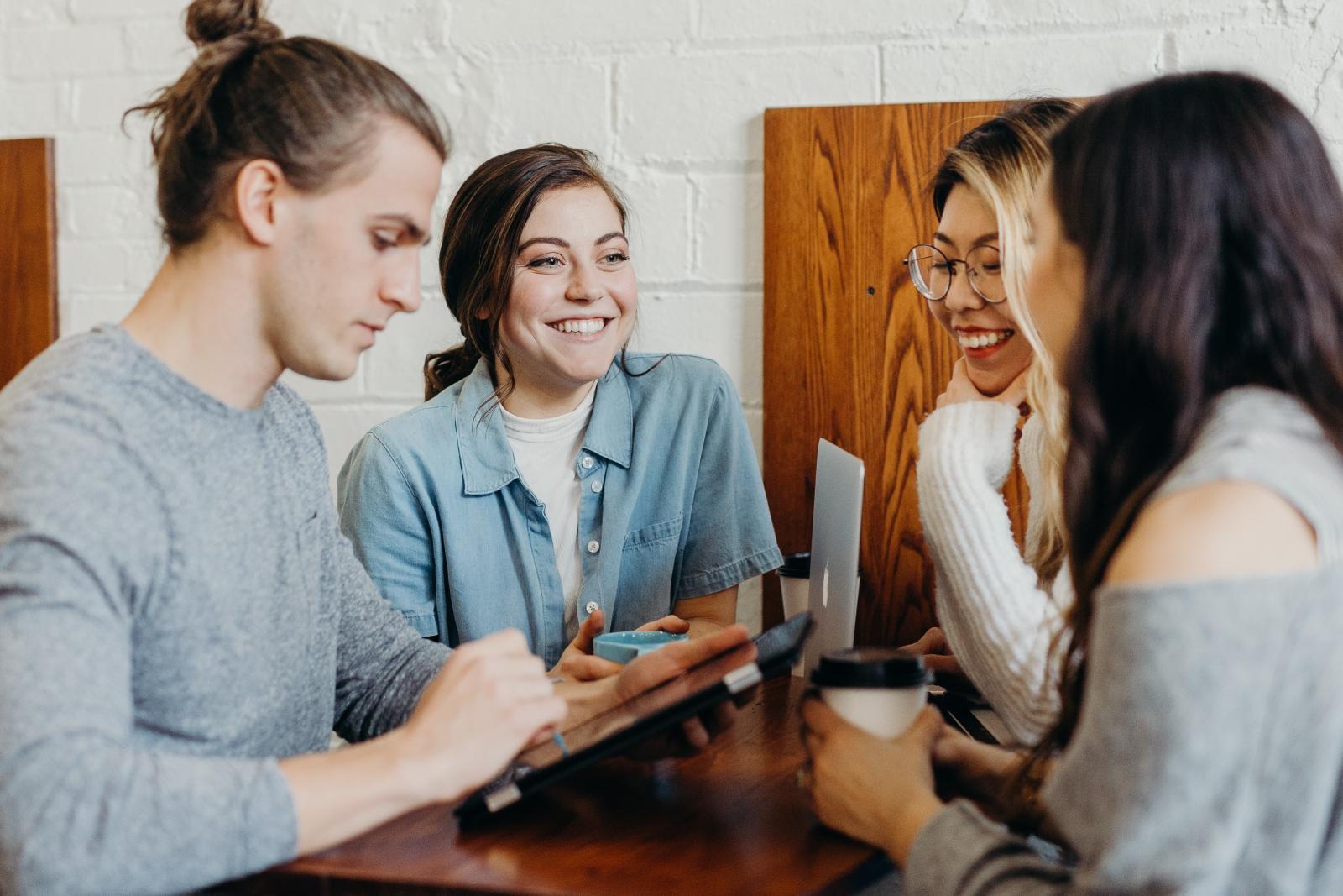 Quattro ragazzi e ragazze che sorridono attorno ad un tavolo, uno usa un tablet, una usa un pc, una afferra un caffè