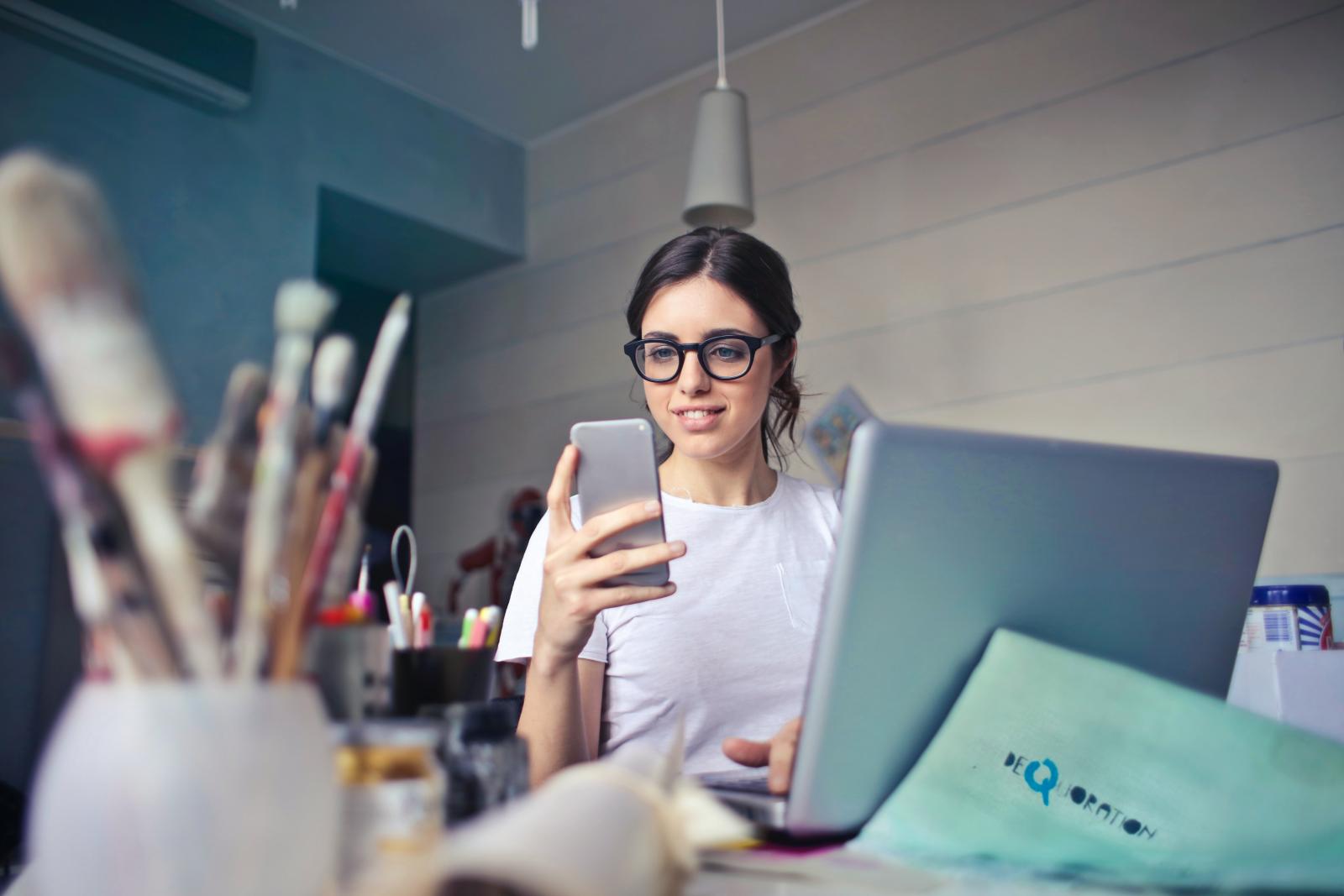 una ragazza guarda il telefono, seduta davanti ad un computer, la scrivania è piena di oggetti (foglio, pennelli, biro)