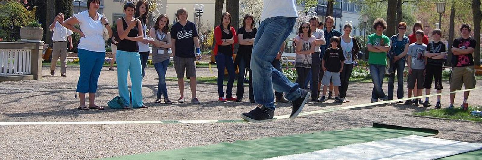 Un ragazzo cammina su una fettuccia sospesa, un pubblico lo guarda