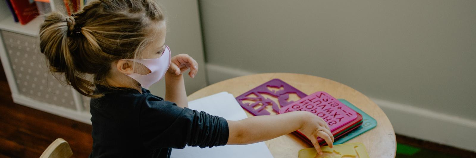 Bambina con la mascherina, seduta ad un tavolo in classe con quaderno e stencil.