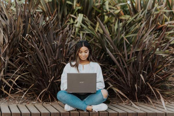 Ragazza seduta per terra che scrive su un pc, sullo sfondo alcuni cespugli