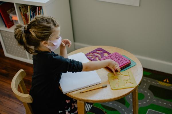 Bambina con la mascherina, seduta ad un tavolo in classe con quaderno e stencil.